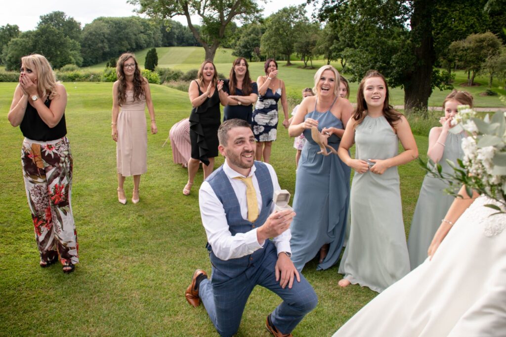 As the bouquet toss gathered the excited guests, laughter and anticipation filled the air. The bride held her flowers high, ready to throw—but instead of launching them into the crowd, she turned and gracefully walked towards her maid of honour. The moment her friend turned around, confusion turned to shock as she saw her partner down on one knee, ring in hand, eyes full of love. Gasps and cheers erupted from the wedding party as the realisation swept over them. Some covered their mouths in surprise, others clapped with joy, while a few wiped away happy tears. The groom-to-be, dressed in a smart blue waistcoat and gold tie, beamed with nervous excitement as he asked the most important question of his life. And then, the magic happened—she said YES! A wave of emotion rippled through the guests as they celebrated this unexpected yet beautifully orchestrated moment, captured in a single, timeless photograph.
