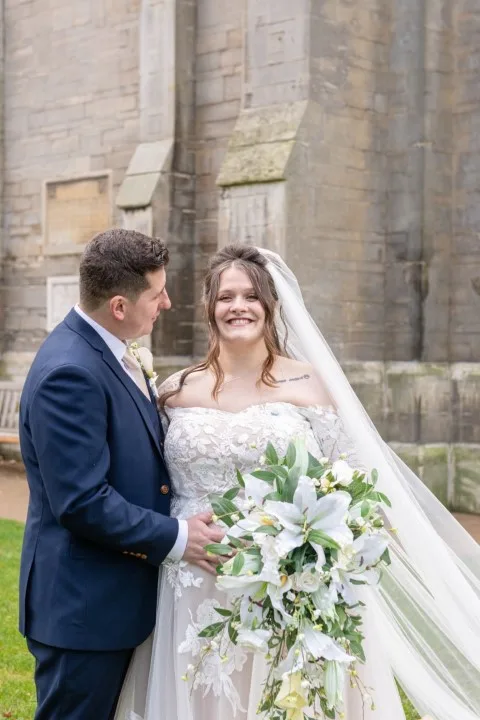 Bride and groom smiling outside a stone church, with the bride holding a large cascading bouquet of white lilies and roses