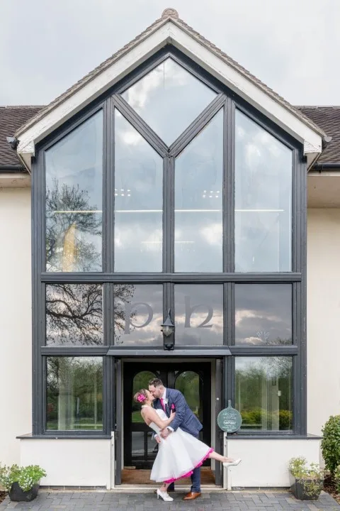 Bride and groom sharing a kiss in front of a modern glass-fronted venue on their wedding day
