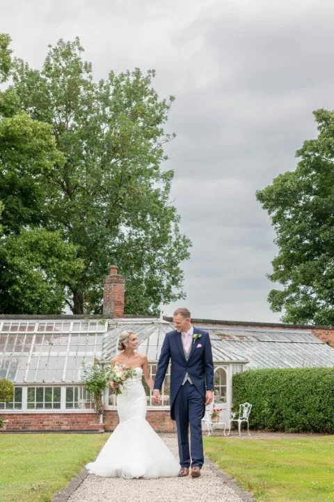 Bride and groom holding hands and smiling while walking past a vintage greenhouse on their wedding day
