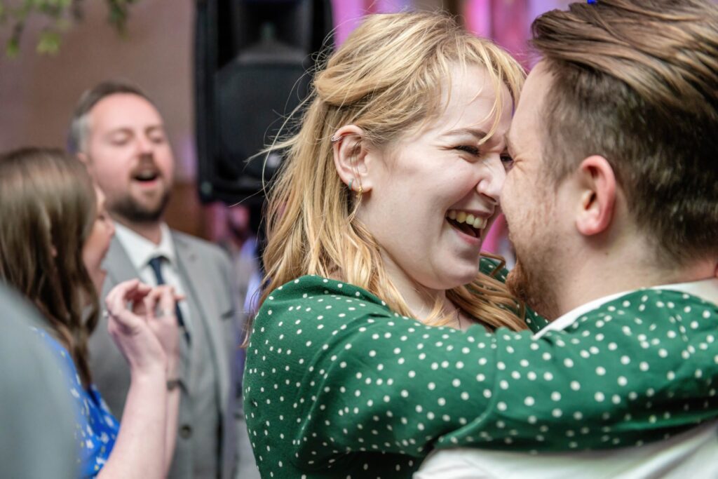 Two people sharing a heartfelt laugh while dancing closely at a social event, with others in the background.