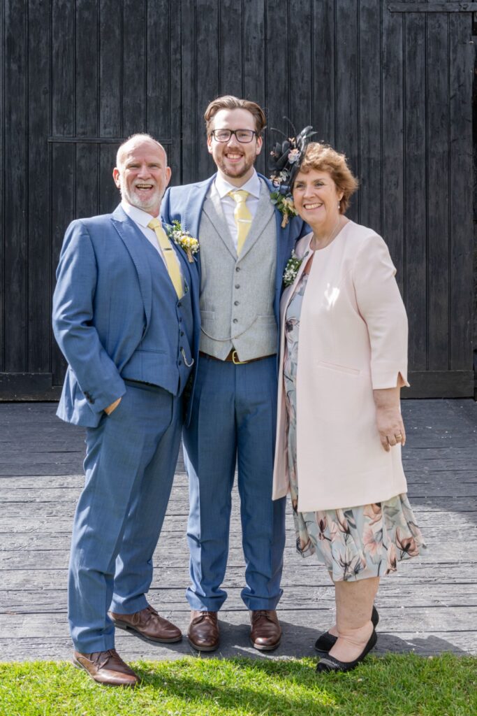 Three people smiling for the camera on a wooden deck, with two men in suits and a woman in a dress and jacket.