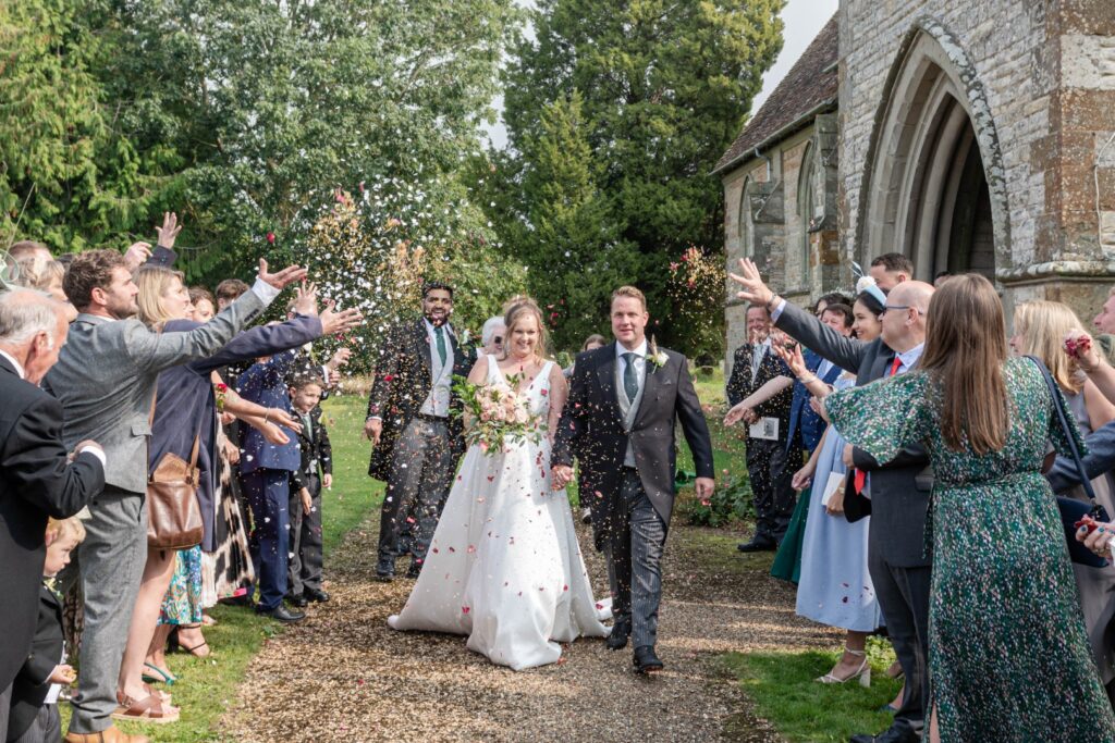 Bride and groom walk through a joyful confetti tunnel outside a countryside church, surrounded by cheering wedding guests – candid and fun wedding moment captured by LB Photography