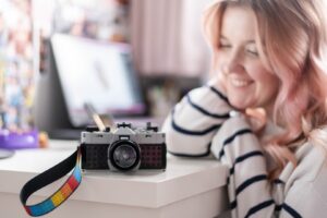 A smiling woman with pink hair sits beside a camera on a white table, with a colorful strap and a blurred background of digital screens.
