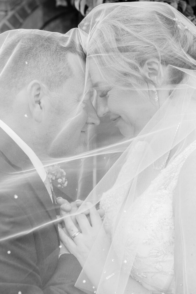Bride and groom smiling with their foreheads together under a flowing veil in black and white wedding portrait