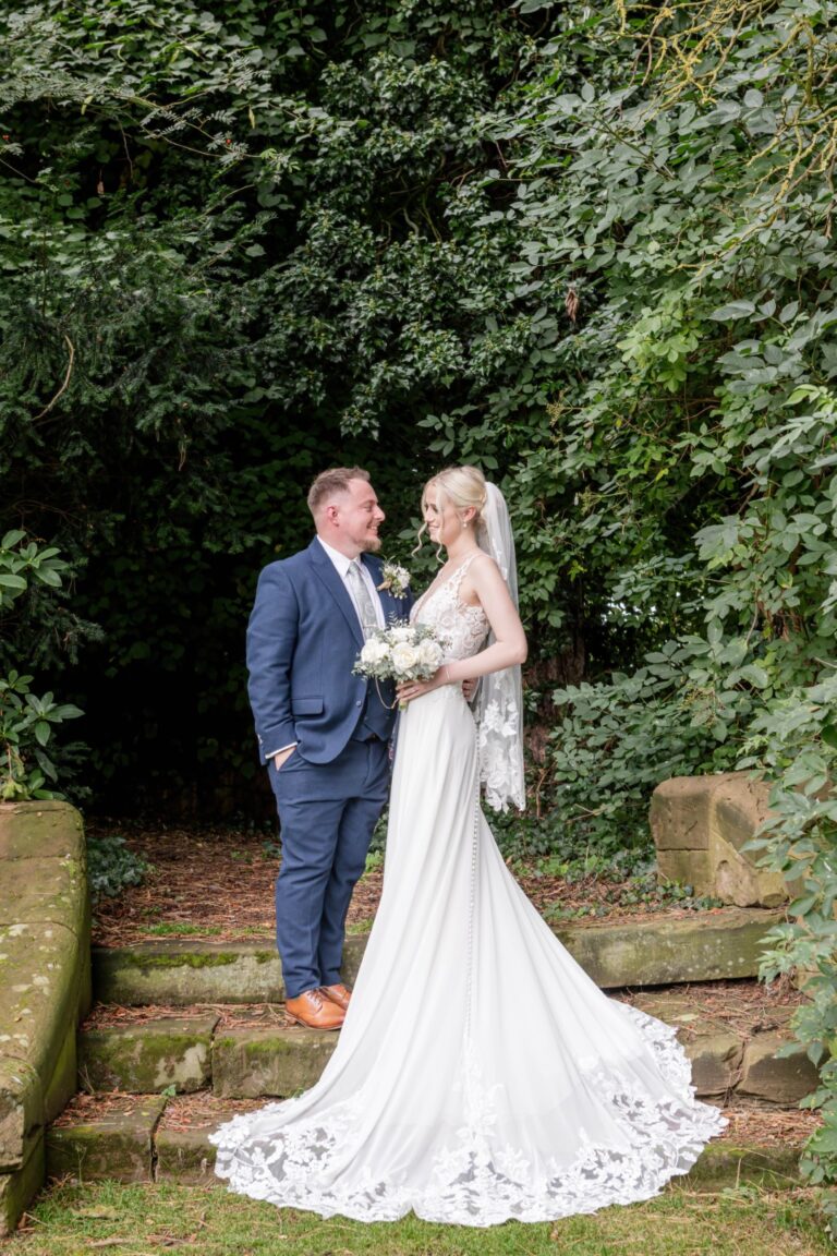 A bride and groom share a joyful moment amidst greenery, with the bride wearing a flowing gown and holding a bouquet of white roses. Elegant 16th-century Weston Hall wedding venue surrounded by Staffordshire countryside