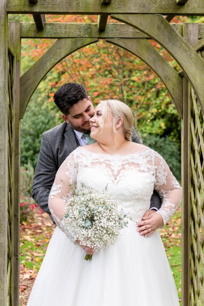 A couple embraces under a wooden arch in a garden, the bride in a lace gown holding a bouquet of flowers, with colorful foliage in the background. Elegant 16th-century Weston Hall wedding venue surrounded by Staffordshire countryside