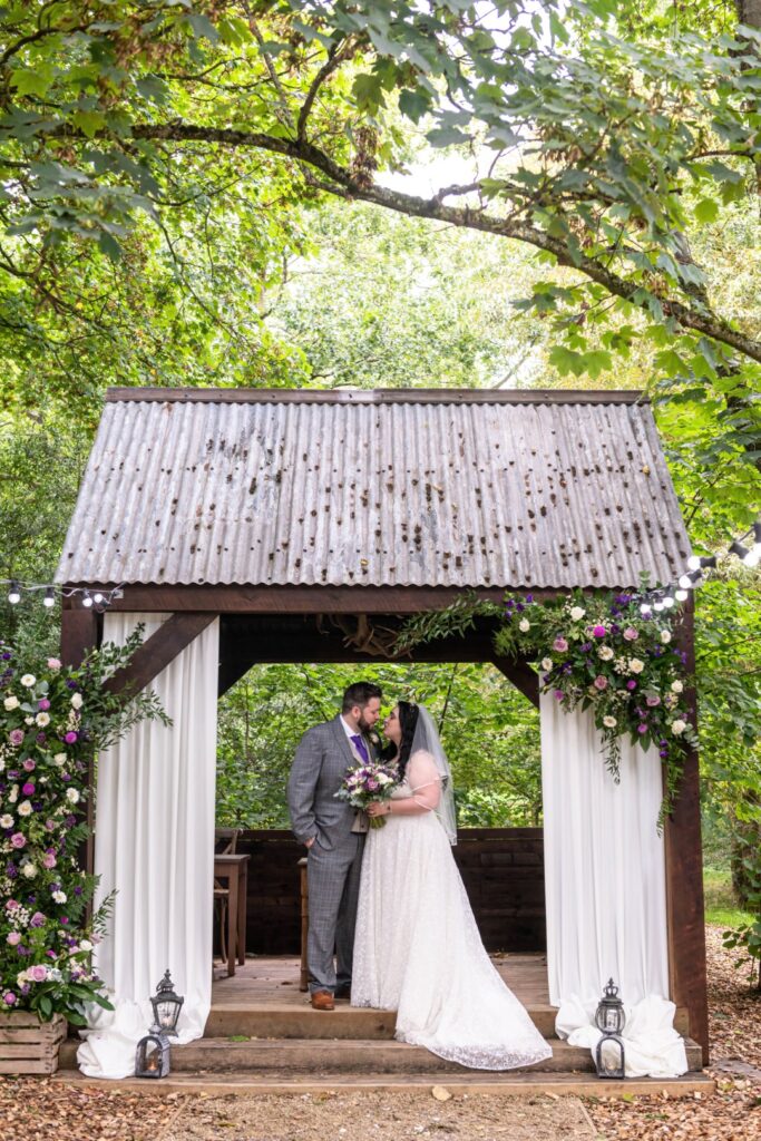 Bride and groom sharing a kiss under a rustic woodland arch decorated with flowers and white drapes during an outdoor wedding ceremony