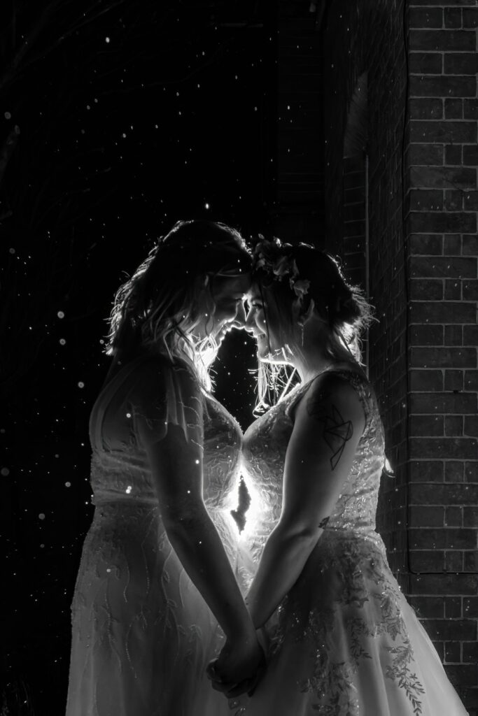 Two brides holding hands under evening light with raindrops falling – romantic and dramatic wedding portrait captured naturally by LB Photography