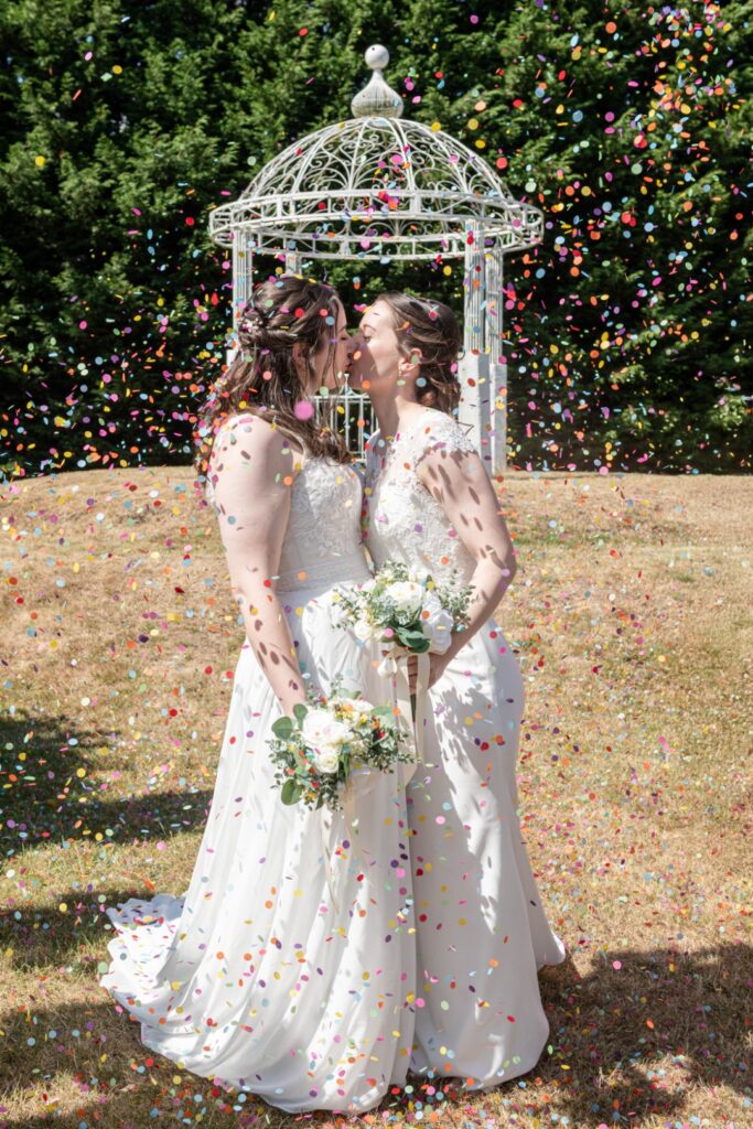 Two brides share a kiss under a shower of colorful confetti, holding bouquets, with a decorative gazebo in the background.