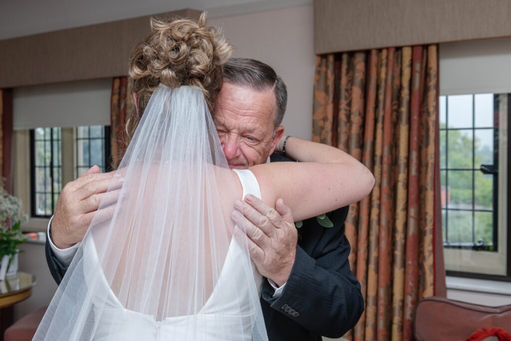 A bride embraces her father in a heartfelt moment, showcasing love and joy before the ceremony, with a soft veil and elegant attire in a warmly decorated room.