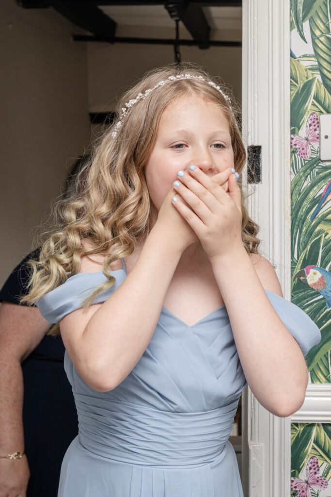 A young girl in a light blue dress looks surprised, covering her mouth with her hands, against a backdrop of vibrant wallpaper.