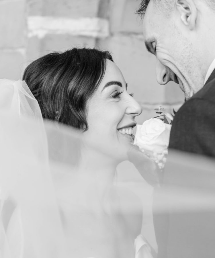 Black and white close-up of a bride and groom smiling at each other, wrapped in the bride’s veil – romantic and natural wedding photography moment